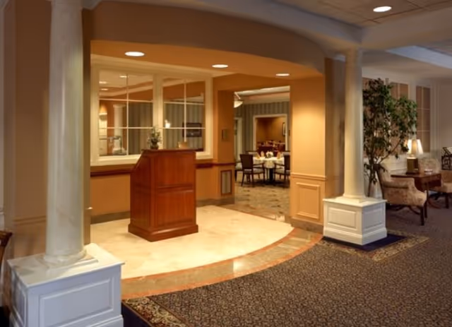Interior view of a senior living facility lobby area with two white columns on either side, a wooden podium in the center, and a glimpse into a dining area with tables and chairs in the background. The space is warmly lit with beige walls and carpeted flooring.