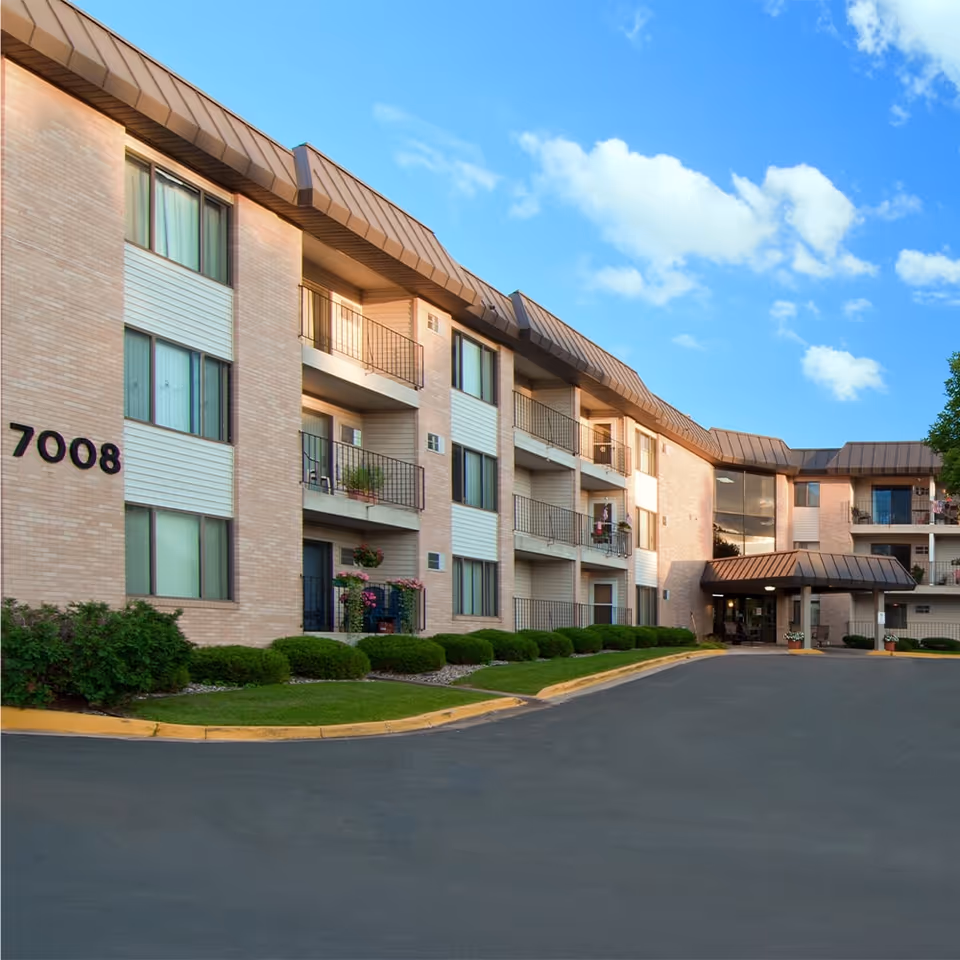Exterior view of a three-story senior living facility building with balconies, large windows, and a covered entrance. The building is surrounded by neatly trimmed bushes and a paved driveway under a blue sky with scattered clouds. The number 7008 is visible on the building's facade.
