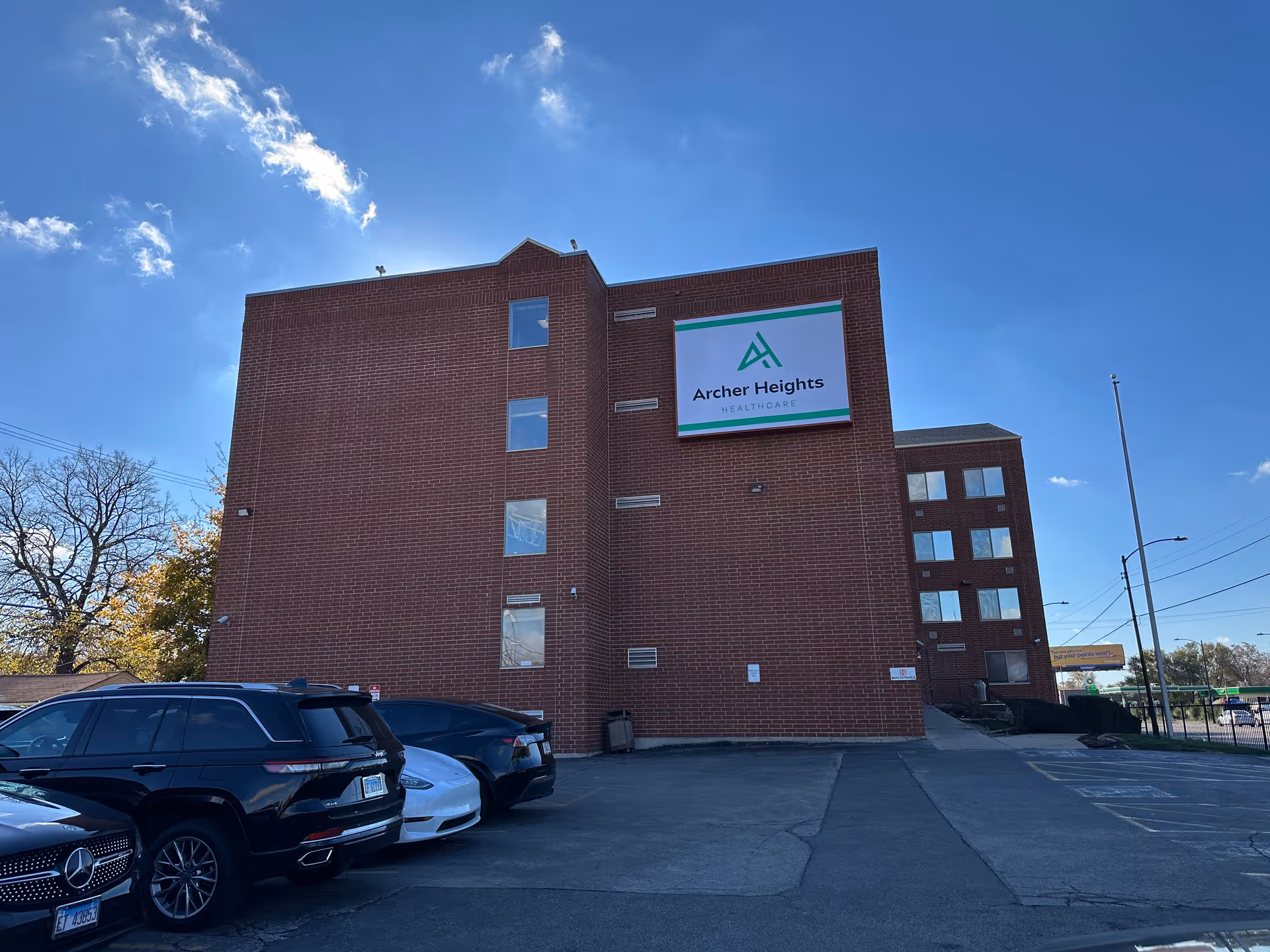Exterior view of a multi-story brick building with a large sign that reads 'Archer Heights Healthcare'. Several cars are parked in front of the building under a clear blue sky with a few clouds.