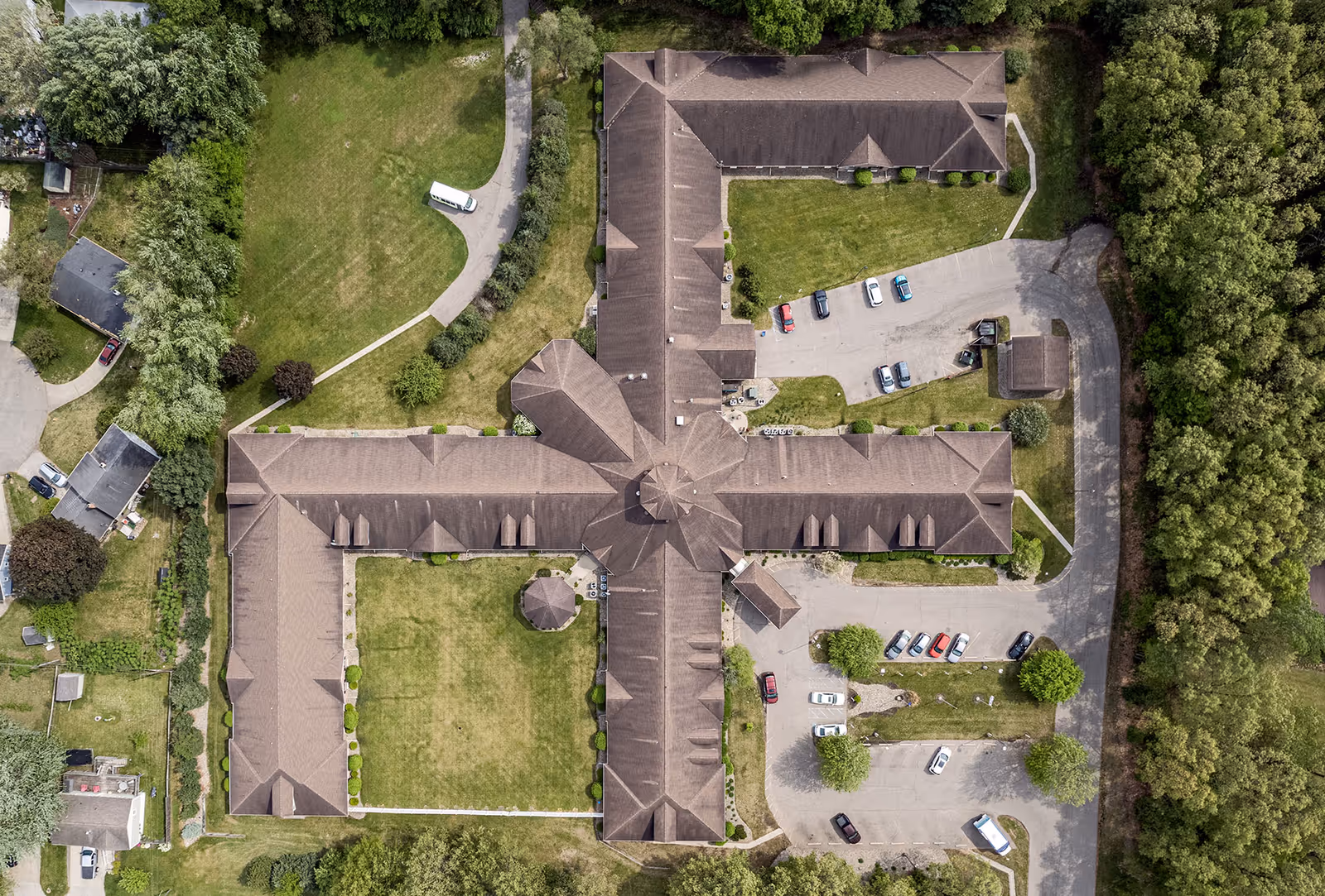 Aerial view of Brentwood at Elkhart Assisted Living facility showing a large building with a cross-shaped layout surrounded by green lawns, trees, a parking lot with several cars, and nearby residential houses.