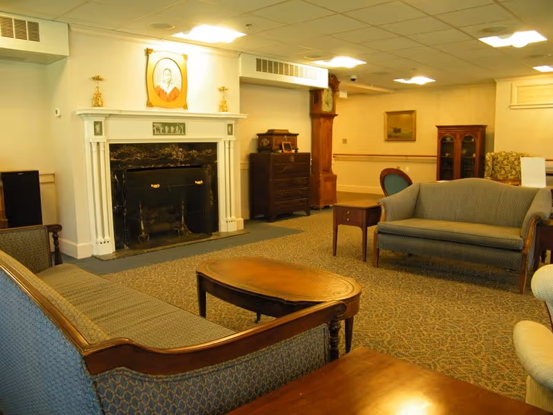 A cozy living room area in a retirement community featuring two upholstered sofas, a wooden coffee table, a fireplace with a white mantel, a grandfather clock, a wooden chest of drawers, and framed artwork on the walls. The room has carpeted flooring and ceiling lights.