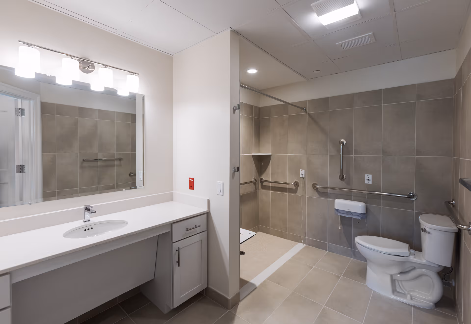 A spacious, modern bathroom with beige tiled walls and floor. The bathroom features a white countertop with an under-mount sink and a large mirror above it, illuminated by four light fixtures. There is an accessible shower area with grab bars and a fold-down seat, as well as a toilet equipped with grab bars for support.