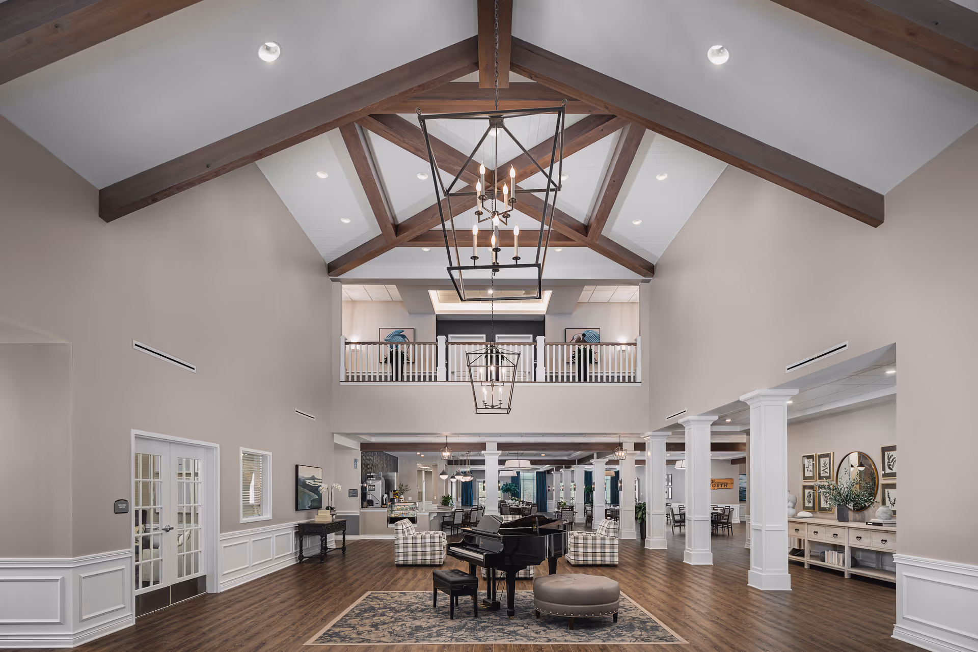 Spacious and elegant interior of a senior living community with high vaulted ceilings featuring exposed wooden beams and modern chandeliers. The room includes a black grand piano on a patterned rug, cushioned chairs, and a seating area. The space has wood flooring, white columns, and a balcony overlooking the main area. Walls are painted in neutral tones with decorative artwork and plants.