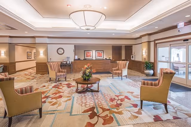 A spacious and well-lit lobby area with a large patterned carpet, four upholstered armchairs arranged around a wooden coffee table with a floral centerpiece, a reception desk in the background, and large windows and glass doors letting in natural light.