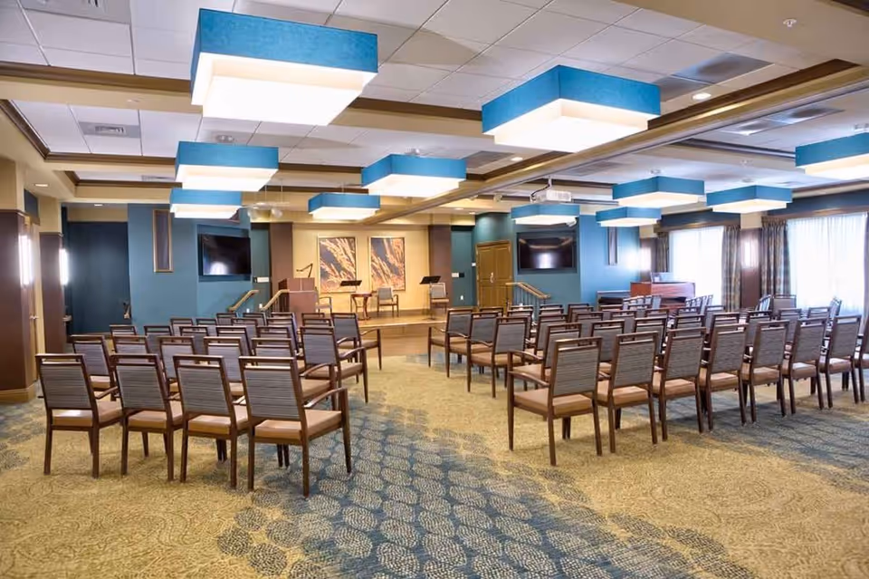 A spacious meeting or event room with rows of wooden chairs facing a small stage area with two chairs and music stands. The room features large blue and white ceiling lights, two wall-mounted televisions, and large windows with curtains allowing natural light to enter.