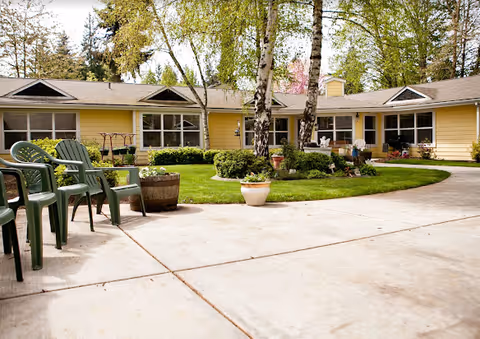 Outdoor courtyard area of Rackleff Place featuring a circular concrete pathway surrounding a grassy area with trees and potted plants. Several green plastic chairs are arranged along the pathway. The building is a single-story structure painted yellow with large windows and a shingled roof.