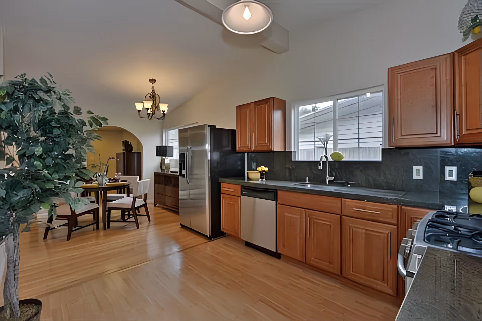 A modern kitchen with wooden cabinets, a stainless steel refrigerator, dishwasher, and stove. There is a window above the sink letting in natural light. The kitchen opens into a dining area with a round table and four chairs, a chandelier overhead, and a large potted plant on the left side.