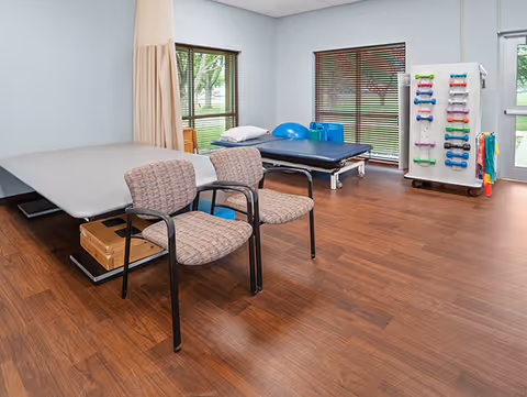 A therapy room with two padded chairs, two therapy tables, exercise equipment including small dumbbells on a rack, and large windows with wooden blinds overlooking a green outdoor area.