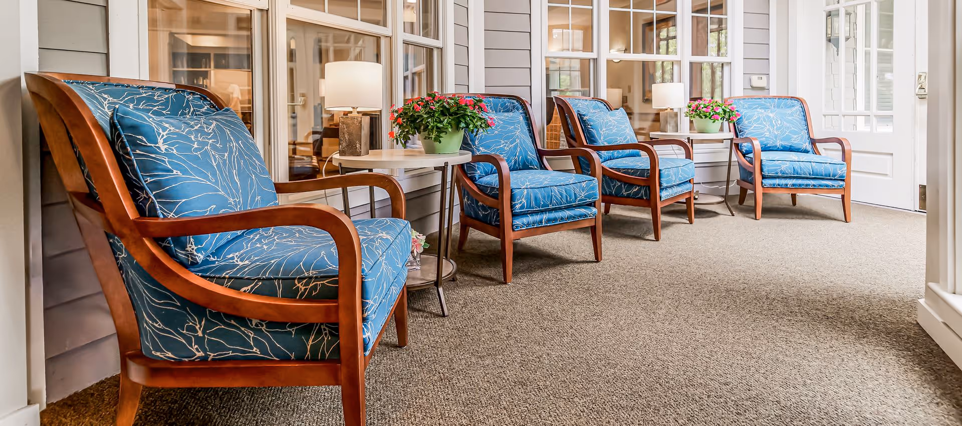 A cozy indoor seating area with four wooden armchairs featuring blue cushions with white branch patterns. Between the chairs are small round tables with potted pink flowers and table lamps. The area has beige carpeting and large windows looking into another room.