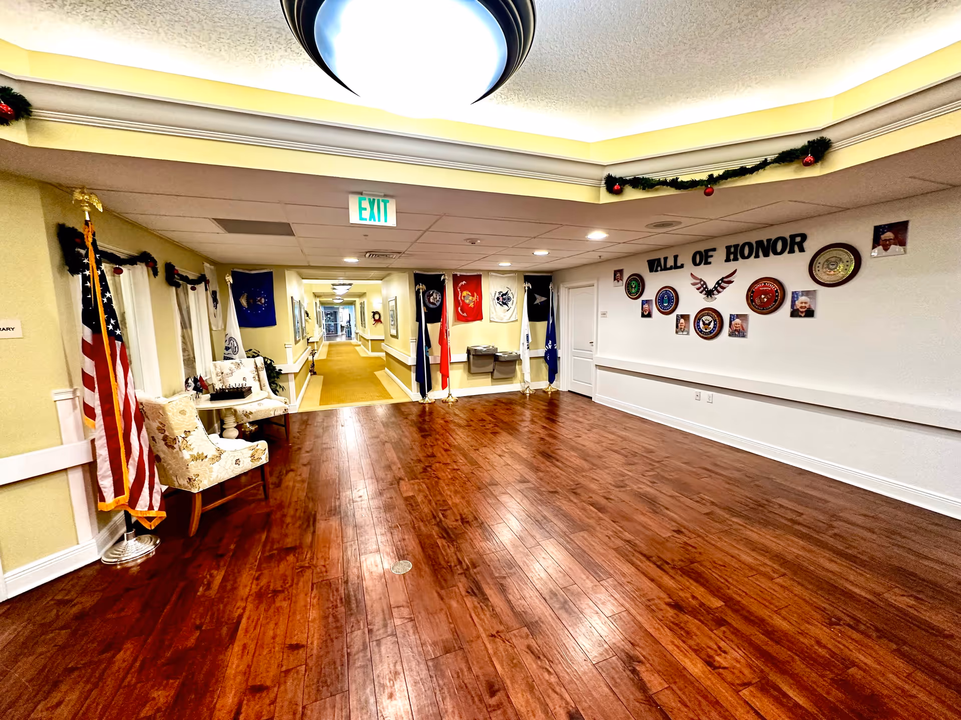 Interior hallway of Lady Lake Senior Living with polished wooden floors, cream-colored walls, and ceiling lights. On the right wall, there is a 'Wall of Honor' display featuring military emblems and photos. Several flags are positioned near the hallway entrance, and two upholstered chairs with a small table are on the left side. The hallway extends into the distance with more lighting and framed pictures along the walls.