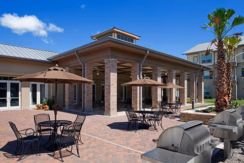 Outdoor patio area with brick pavers, round tables and umbrellas, a covered walkway supported by brick columns, and barbecue grills under a blue sky.