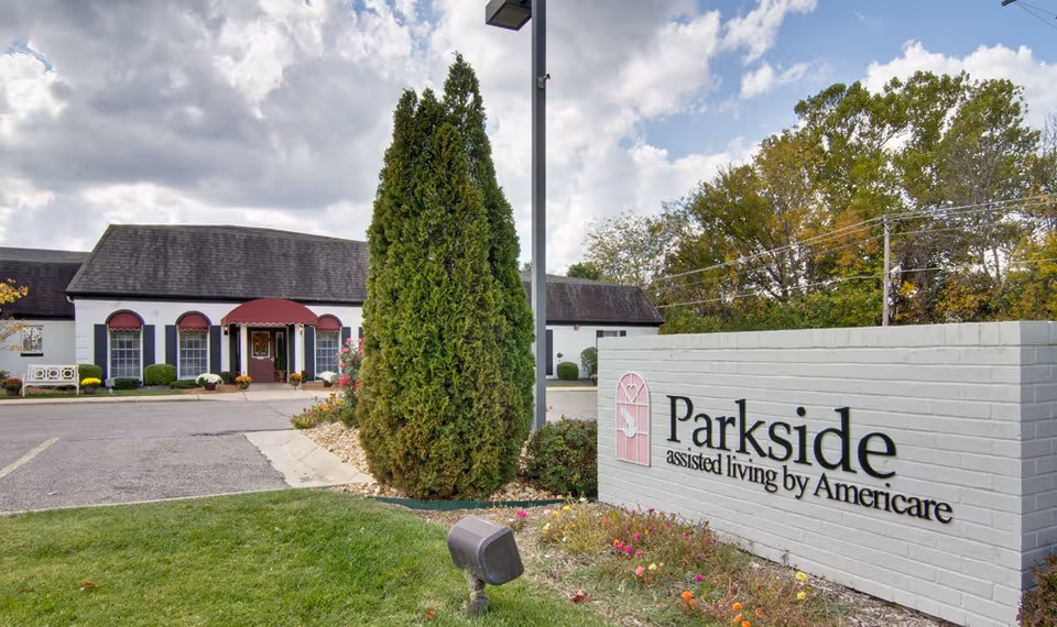Exterior view of an assisted living facility named Parkside, showing a white brick sign with the facility's name and logo, a tall evergreen tree, a lamp post, and a building with a dark roof and red awnings over the windows and entrance. The sky is partly cloudy and there are trees in the background.