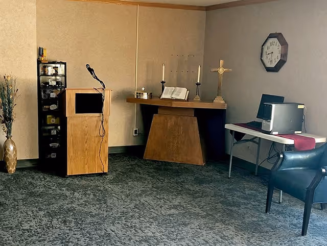 A small room with a wooden podium equipped with a microphone, a table holding a cross, two candles, and an open book, a clock on the wall, a computer on a folding table with a red runner, a black chair, and a tall vase with decorative dried plants in the corner.