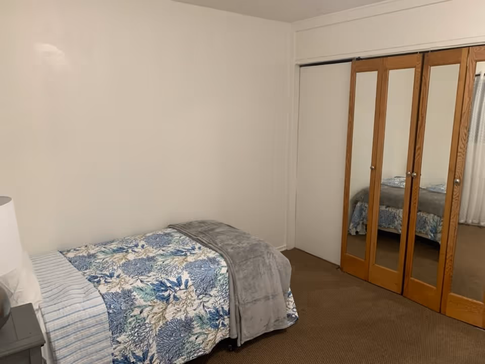 Small bedroom with a single bed covered in blue floral bedding, a gray throw, and mirrored closet doors against a neutral wall.