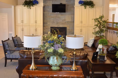 A cozy living room area featuring two brown leather sofas, two brass table lamps with white shades on a wooden table, a large floral arrangement in a green vase, and a stone fireplace with a mounted flat-screen TV above it. The room also includes cream-colored cabinets on either side of the fireplace, two striped armchairs, and various green plants adding a touch of nature.
