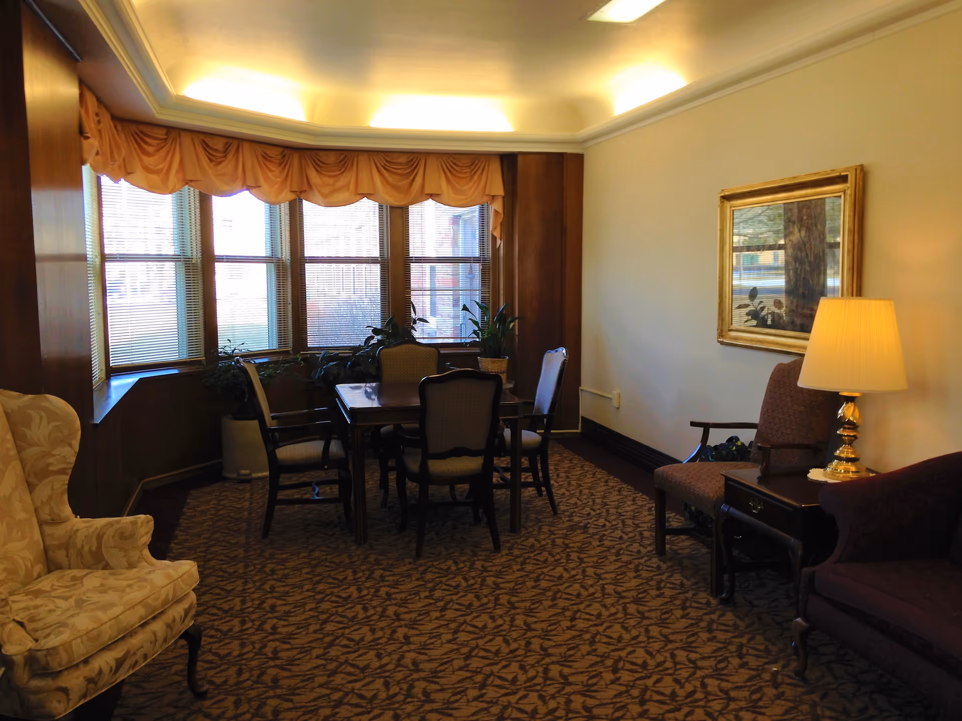 A cozy sitting area in an assisted living facility with a patterned carpet, a wooden table surrounded by four chairs near a large window with blinds and a valance. There are several potted plants on the window ledge. To the right, there is a side table with a lamp, an armchair, and a framed mirror on the wall. Another upholstered armchair is visible on the left side of the image.