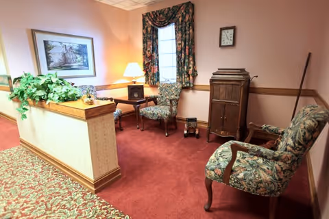 A cozy sitting area with floral upholstered chairs, a small wooden table with a lamp, a framed picture on the wall, a clock, and a wooden cabinet. The room has red carpet flooring and floral curtains on the window.