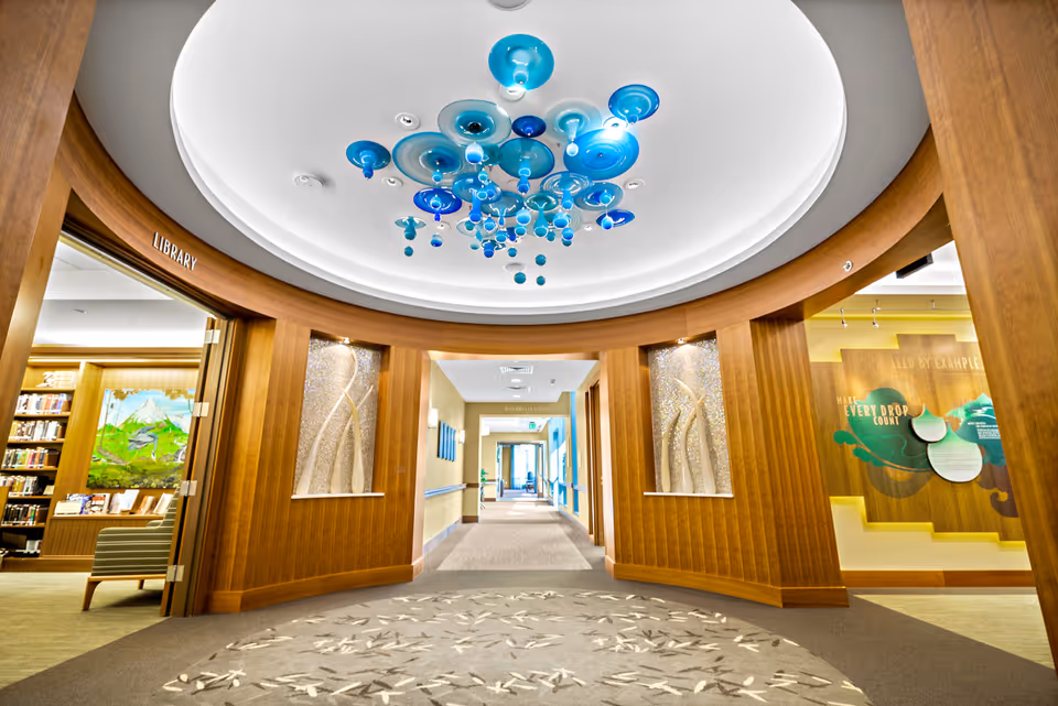 Bright wood-trimmed interior lobby corridor with a blue glass ceiling installation and an entrance to the library.