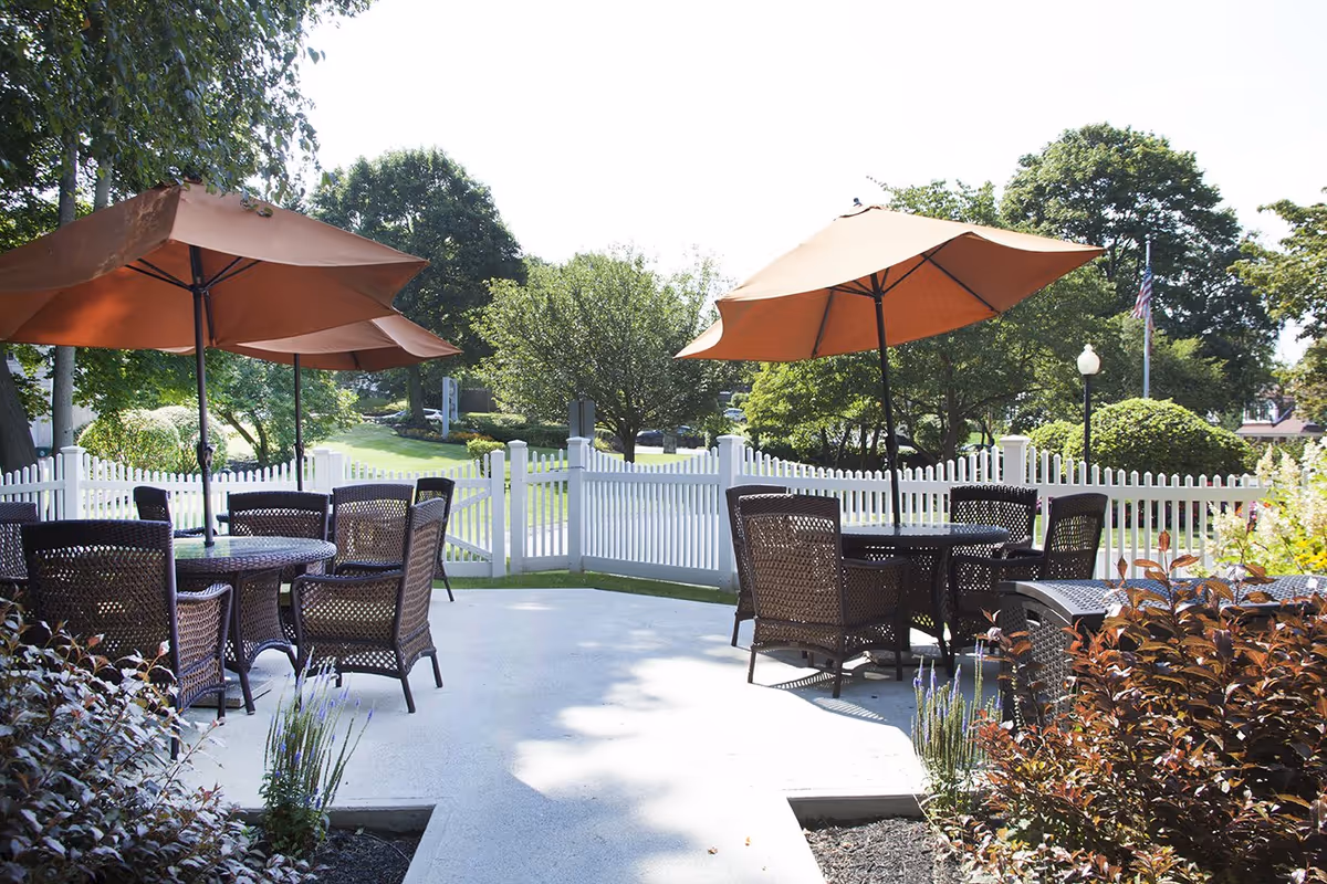 Patio with wicker tables and orange umbrellas overlooking a white picket fence and landscaped grounds.