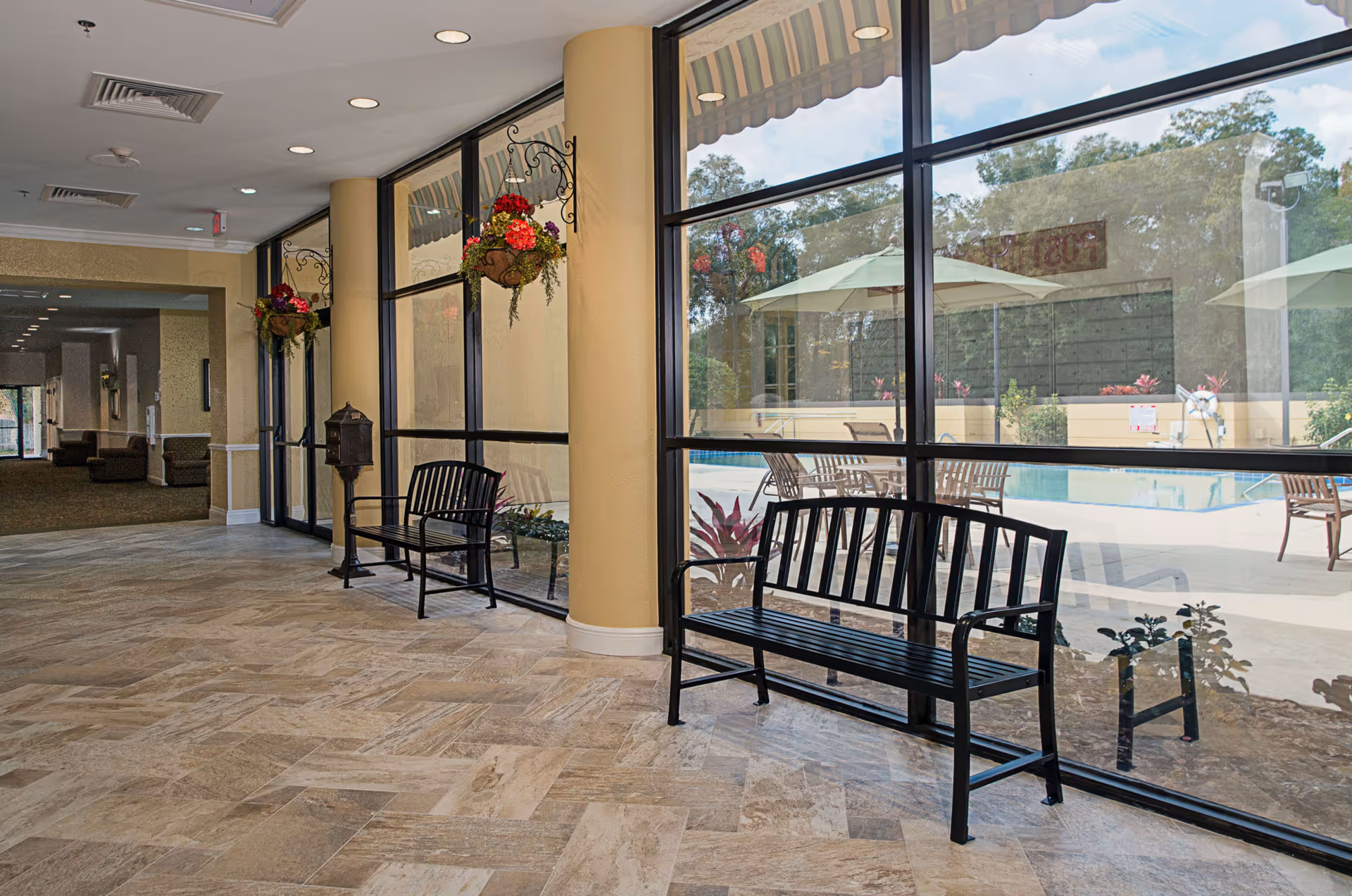 Indoor hallway area with large floor-to-ceiling windows overlooking an outdoor pool area with tables, chairs, and umbrellas. The hallway has tiled flooring, two black metal benches, hanging flower baskets, and beige walls with columns.
