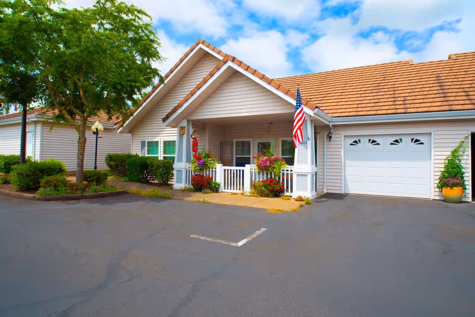 Exterior view of a single-story residential building with beige siding and a brown shingled roof. The entrance features a small covered porch decorated with hanging flower baskets and an American flag. There is a garage door to the right and a tree with surrounding greenery to the left. The sky is partly cloudy.