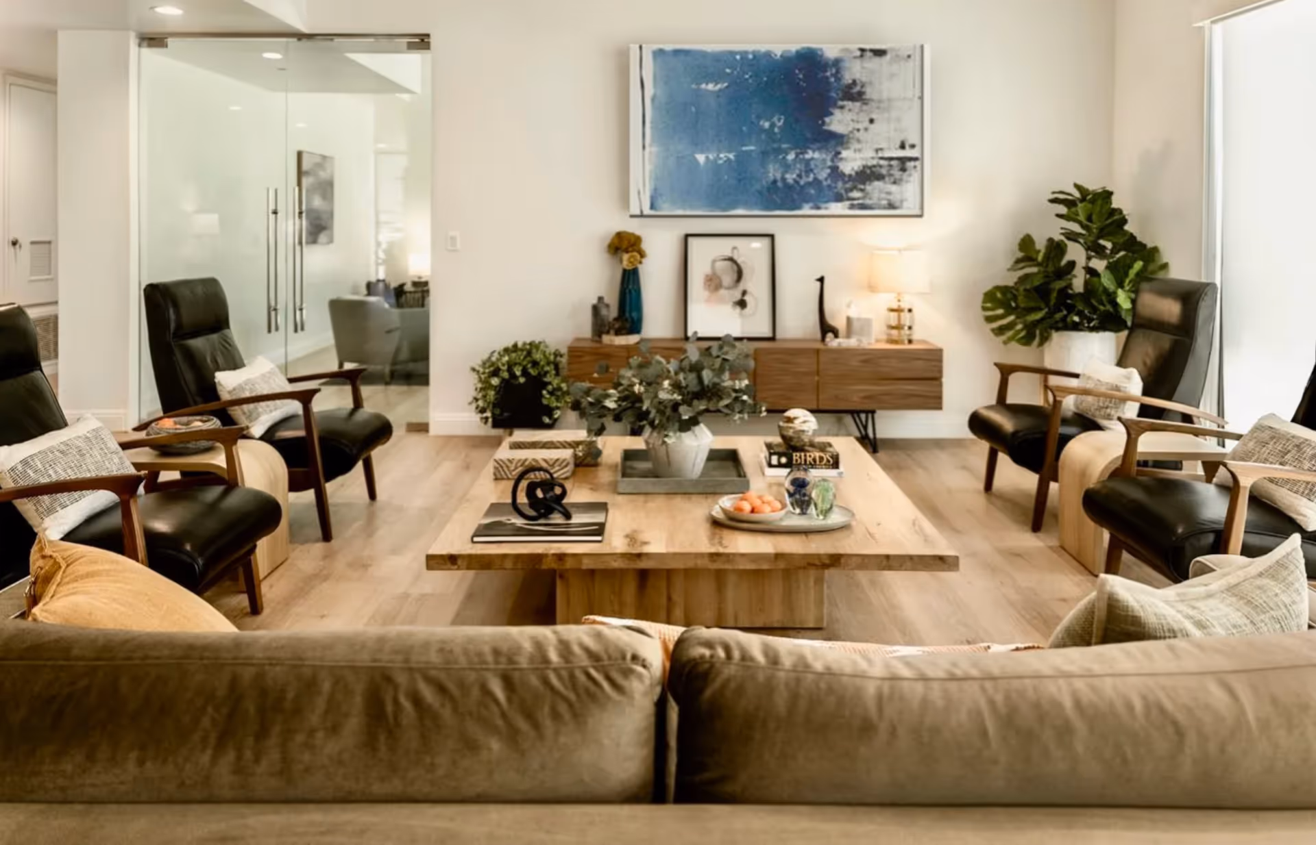 Bright communal living room featuring a sofa facing a wooden coffee table surrounded by black leather armchairs and abstract wall art.