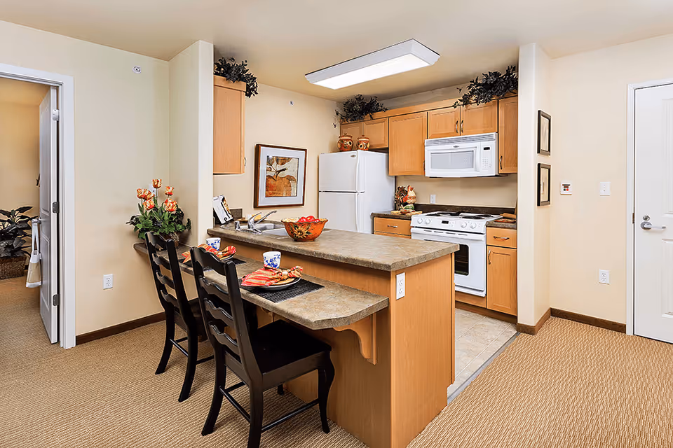 Open kitchen with a breakfast bar and three stools, refrigerator, stove, microwave, and wooden cabinets in a senior living apartment.