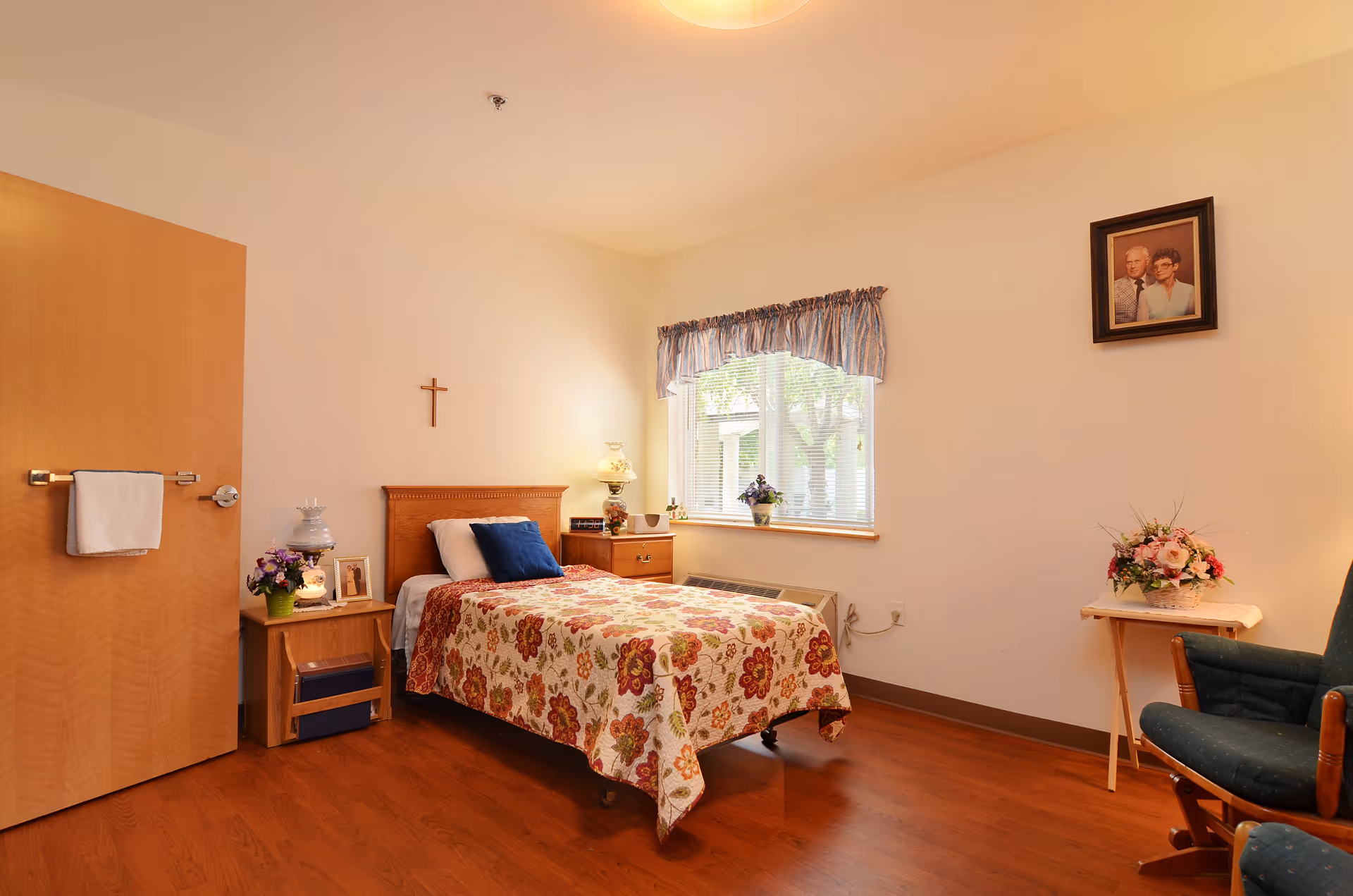 A cozy bedroom with a single bed covered in a floral quilt, a wooden headboard, and a blue pillow. There is a wooden nightstand with a lamp, flowers, and a framed photo next to the bed. A window with blinds and a valance lets in natural light. A wooden door with a towel hanging on the handle is on the left. On the right side, there is a small table with a flower arrangement and a green upholstered chair. A framed portrait hangs on the wall above the chair.