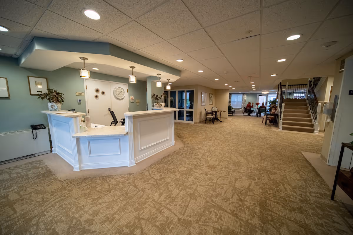 Spacious interior lobby area of a senior living facility with a white reception desk on the left, beige patterned carpet, recessed ceiling lights, and a seating area with people in the background near large windows. A staircase is visible on the right side.