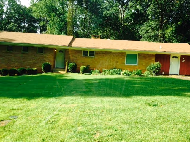 Single-story brick building with a tan roof surrounded by green grass and trees in the background under a clear sky.