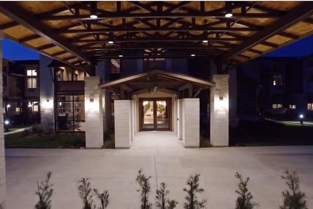 Night view of the entrance to a building with a covered walkway supported by stone pillars and wooden beams. The entrance has glass doors and is illuminated by wall-mounted lights. Small shrubs are visible in the foreground.
