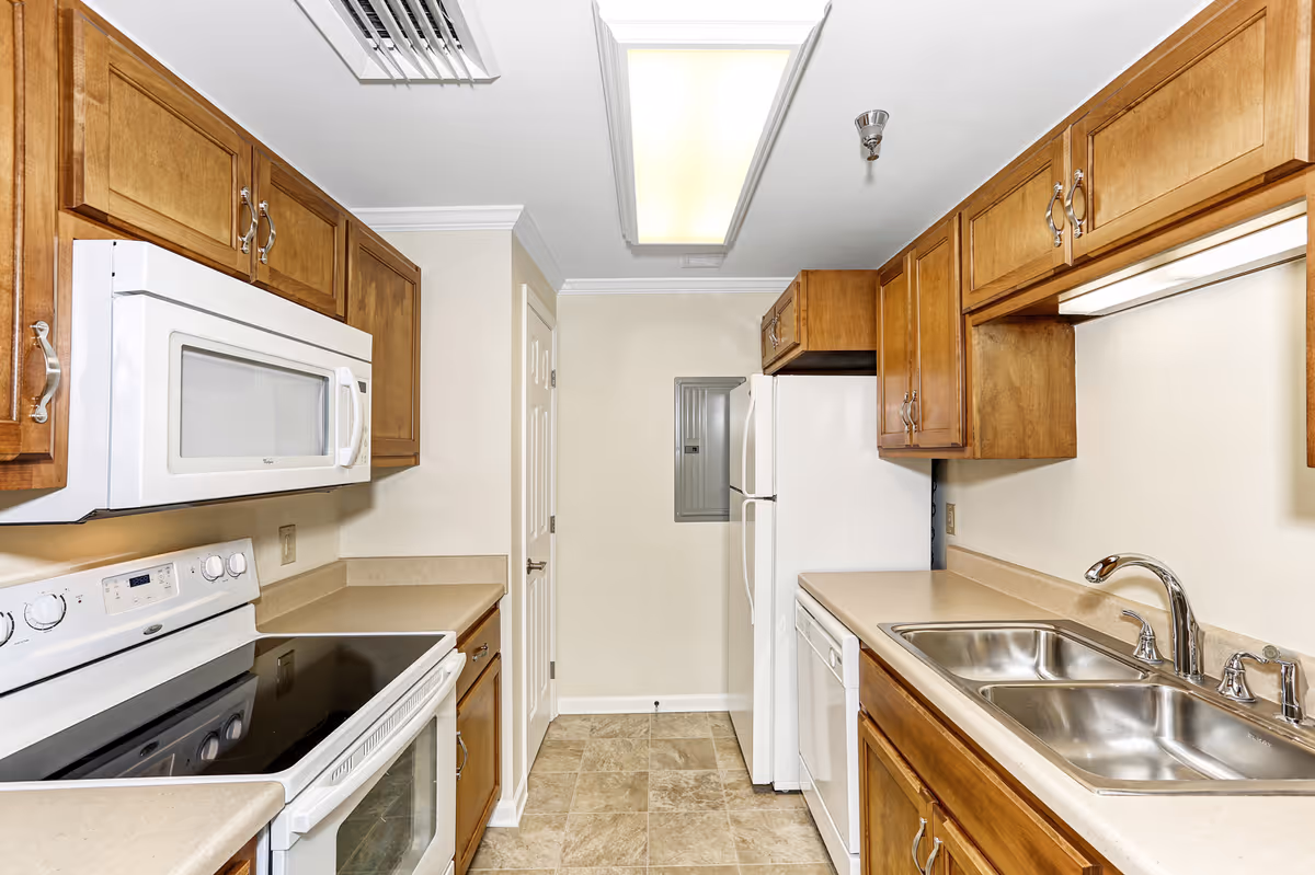 A clean, well-lit kitchen with wooden cabinets, a white microwave above a white electric stove, a double stainless steel sink with a chrome faucet, a white dishwasher, and a white refrigerator. The floor is tiled, and there is a fluorescent light fixture on the ceiling.