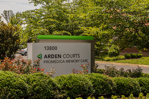 Outdoor sign reading 'ARDEN COURTS PROMEDICA MEMORY CARE' surrounded by shrubs and trees.