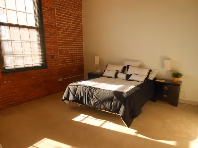 A bedroom with a bed covered in black and white bedding, two bedside tables with lamps, a small plant, and a large window with blinds letting in sunlight. The room has a brick accent wall and beige carpet flooring.