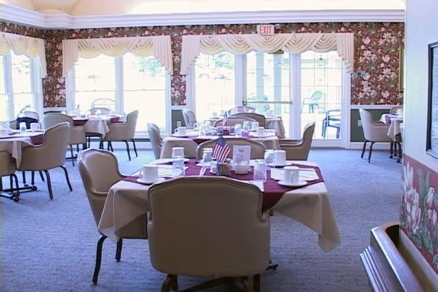 A dining room with several round tables covered with white tablecloths and burgundy overlays. Each table is set with white cups, glasses, plates, and silverware. The room has floral wallpaper, large windows letting in natural light, and beige cushioned chairs around the tables. An American flag is placed on one of the tables.