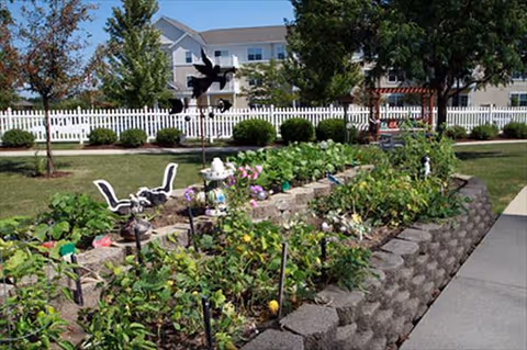 Outdoor garden area with raised flower beds, stone wall, chairs, and a white fence near buildings.