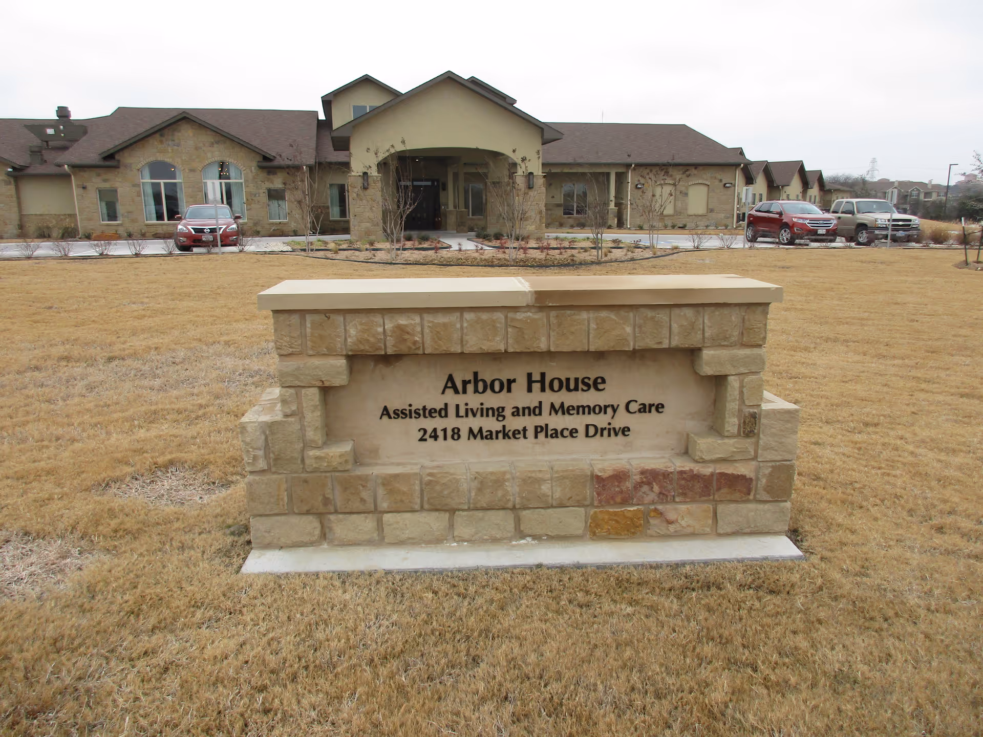 Stone sign in front of a large building with a driveway and parked cars. The sign reads 'Arbor House Assisted Living and Memory Care 2418 Market Place Drive'. The building has a beige exterior with stone accents and a covered entrance.