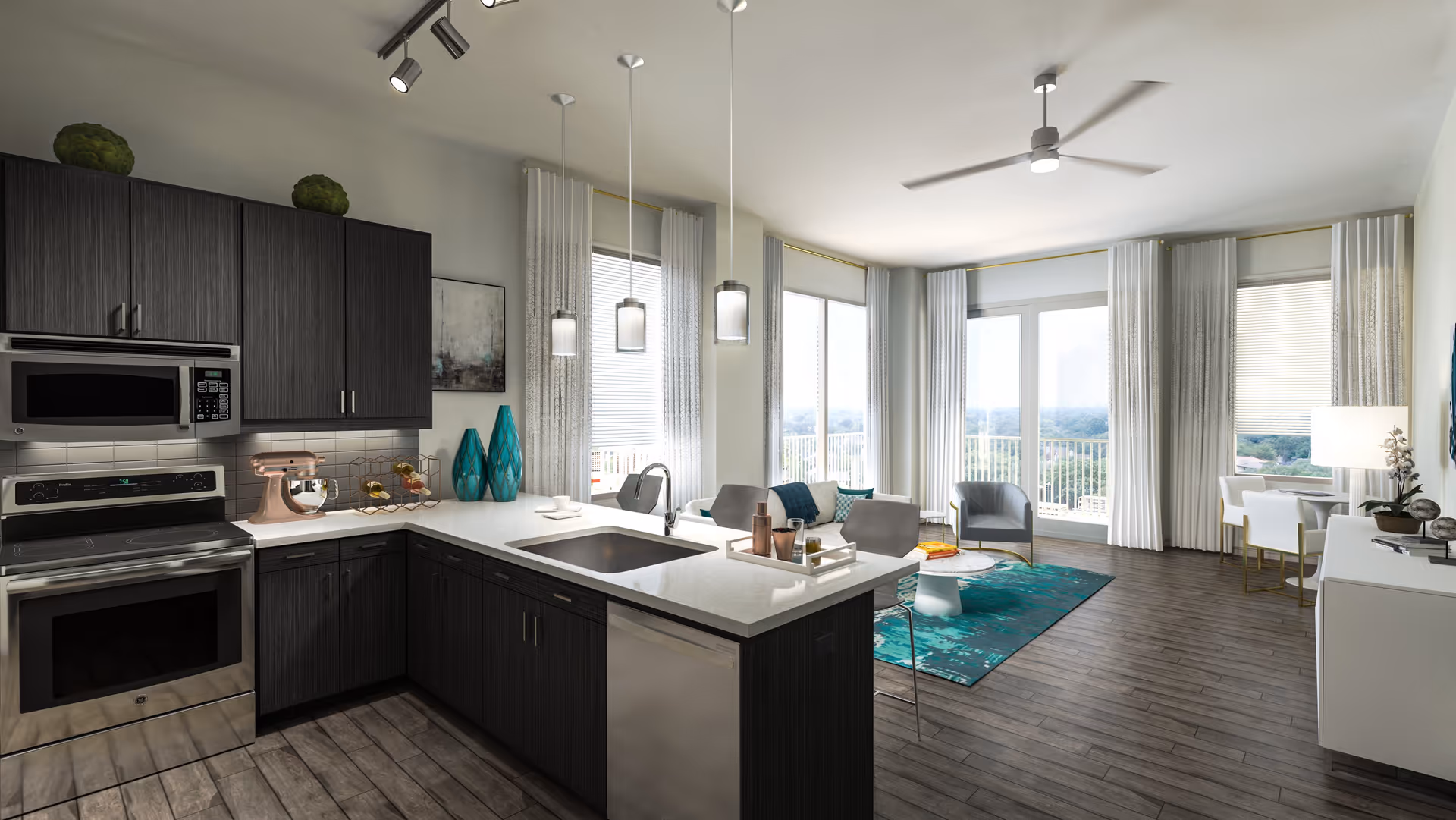 Modern open-concept living space featuring a kitchen with dark wood cabinets, stainless steel appliances, and a white countertop island with a sink. The living area includes a teal rug, gray chairs, a small white coffee table, and large windows with white curtains allowing natural light to fill the room. A dining area with a white table and chairs is visible near the windows.
