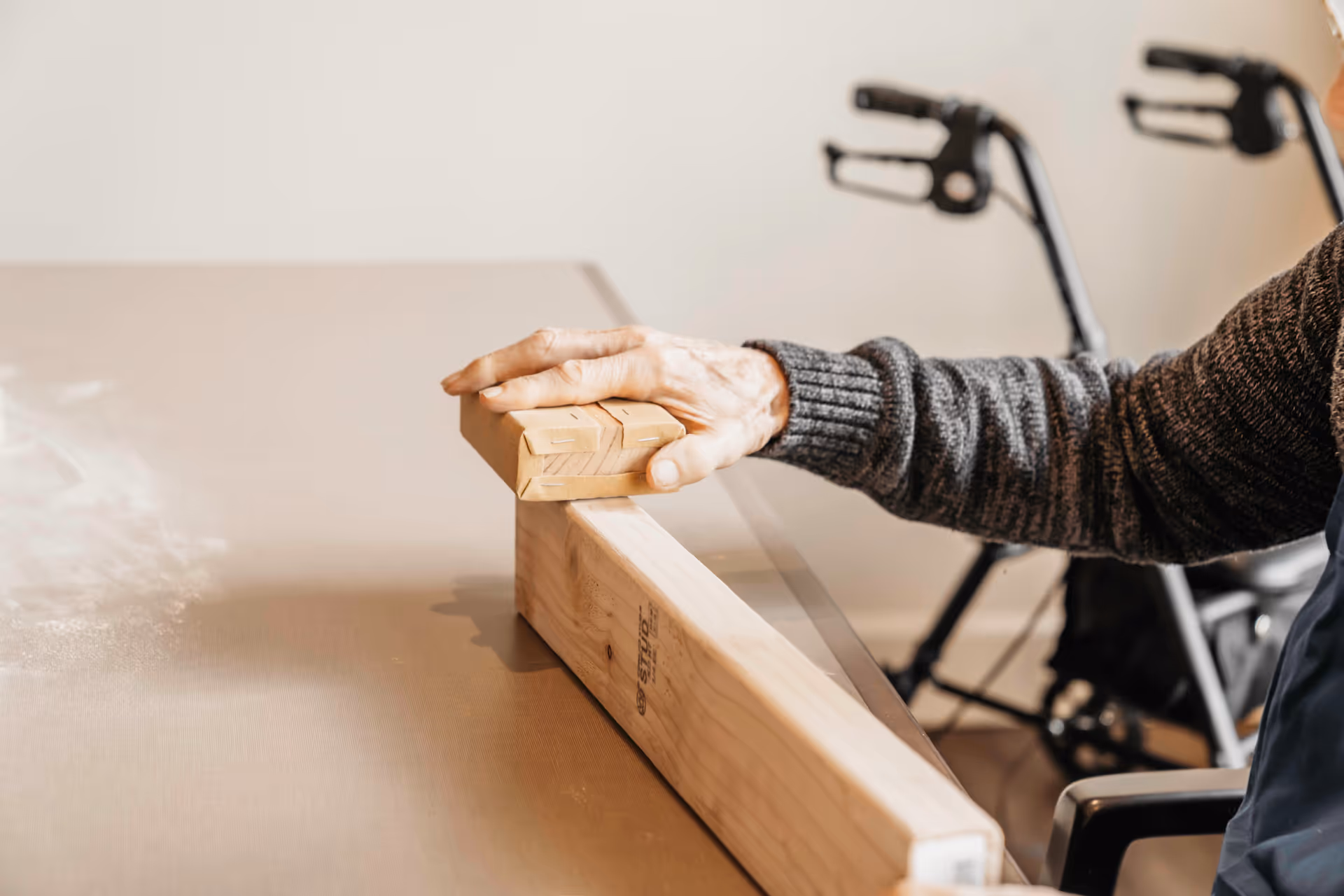 An elderly person wearing a gray sweater is playing a tabletop shuffleboard game, pushing a wooden puck along the smooth surface. A walker is visible in the background.