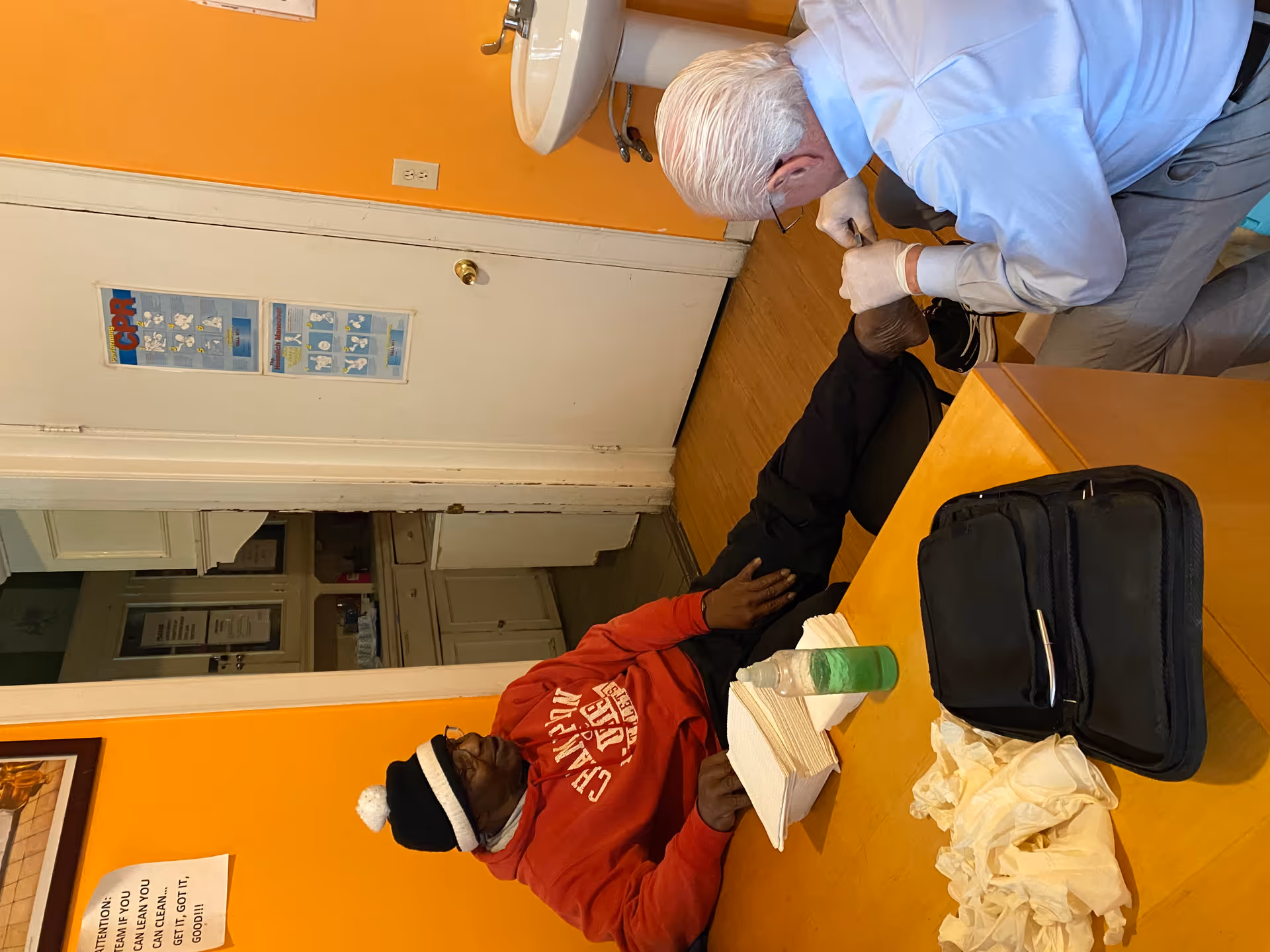 An elderly man wearing a red hoodie and black hat sits with his leg extended on a chair while another man with white hair and glasses, wearing gloves, tends to his foot in a room with orange walls, a white sink, and a door with CPR and Heimlich maneuver posters.