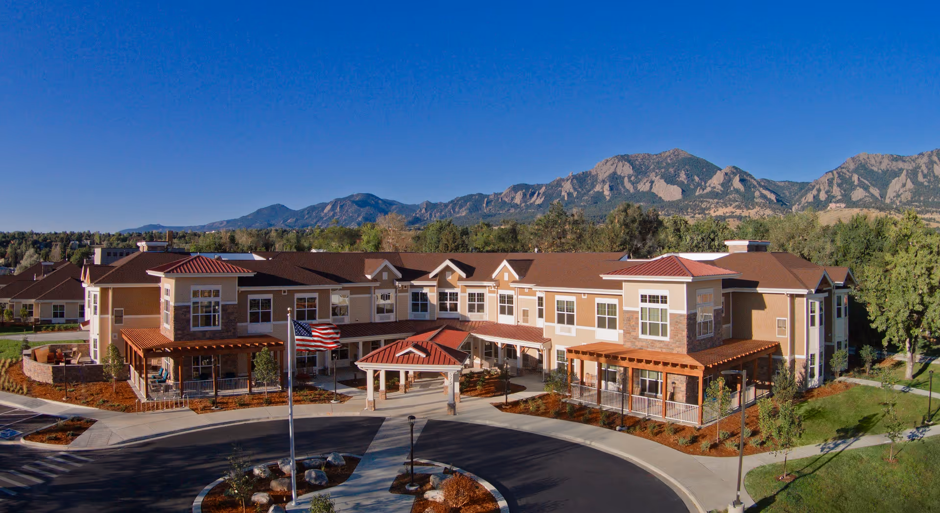 Exterior view of MorningStar Assisted Living & Memory Care of Boulder facility with a two-story building featuring beige and brown tones, multiple windows, covered entrances, and a circular driveway with an American flag in front. The background shows a clear blue sky and mountain range.