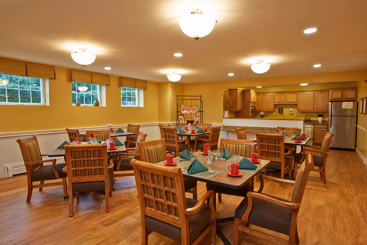 A warm and inviting dining room with several wooden tables and chairs arranged neatly. Each table is set with green folded napkins, red cups, glasses, and silverware. The room has yellow walls, wooden flooring, and three small windows letting in natural light. In the background, there is a kitchen area with wooden cabinets, a stainless steel refrigerator, and a counter with a white front panel. Ceiling lights illuminate the space evenly.