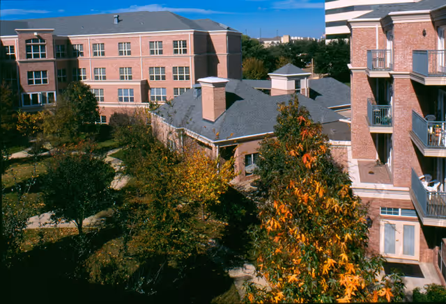 View of a senior living facility courtyard with brick buildings surrounding a landscaped garden area featuring trees with autumn foliage and walking paths under a clear blue sky.