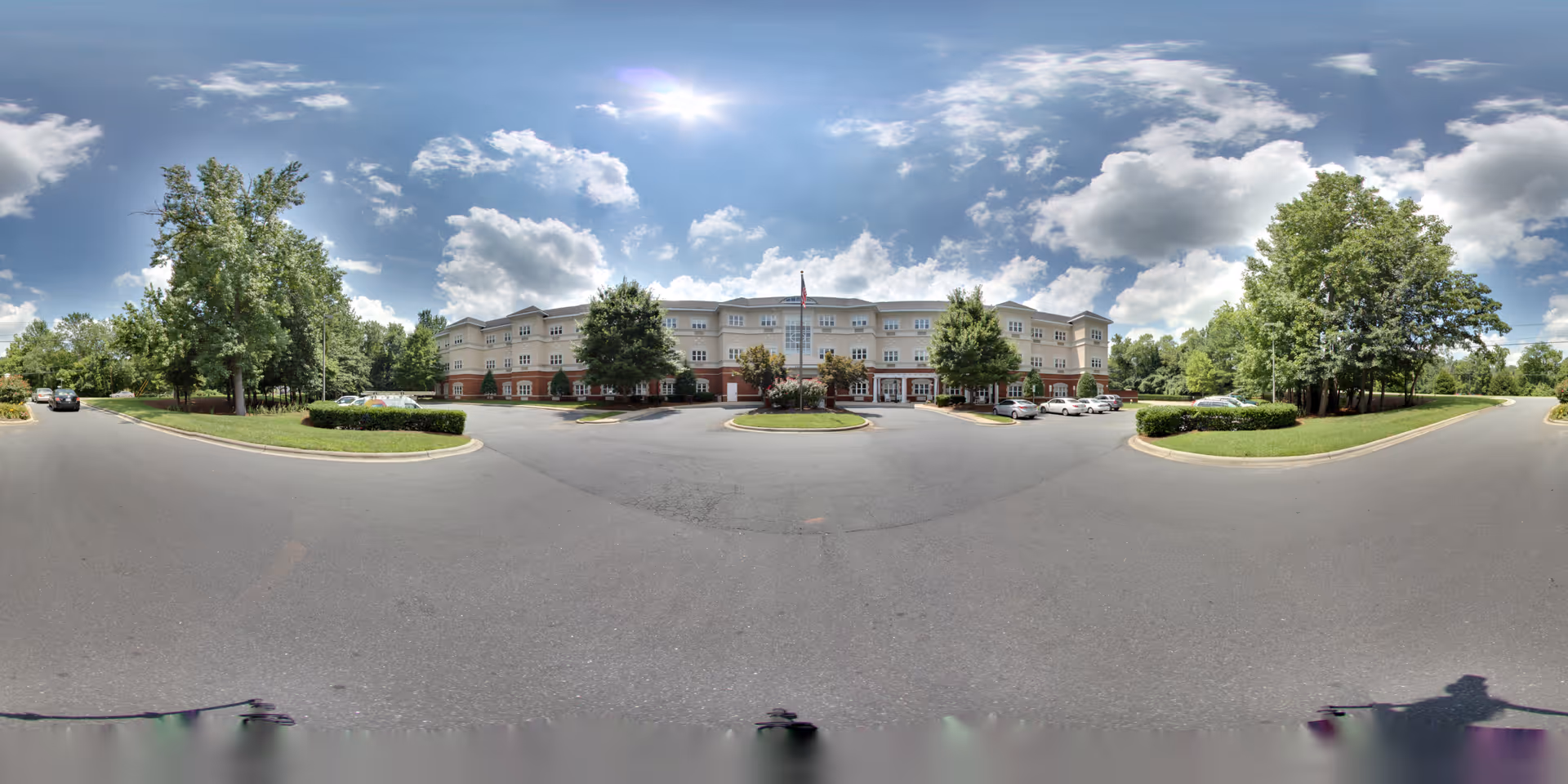 Front view of a three-story senior living building with a circular driveway, flagpole, trees, and parked cars under a partly cloudy sky.