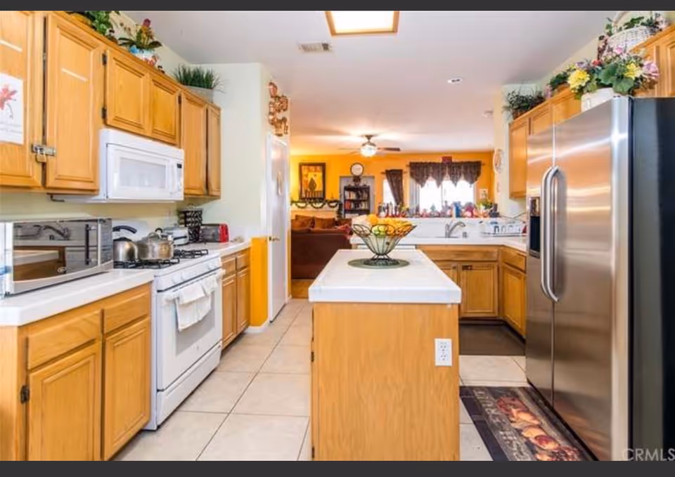 Bright kitchen with wooden cabinets, a center island, white stove and microwave, and a stainless refrigerator opening into a living area.
