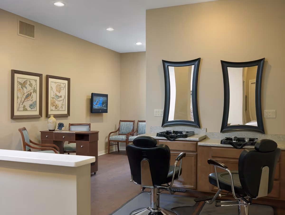 Interior view of a salon area in a senior living facility with two black salon chairs in front of two sinks and mirrors. In the background, there is a small desk with a chair, two framed pictures on the wall, and two additional chairs. The walls are painted beige and the floor is wooden.