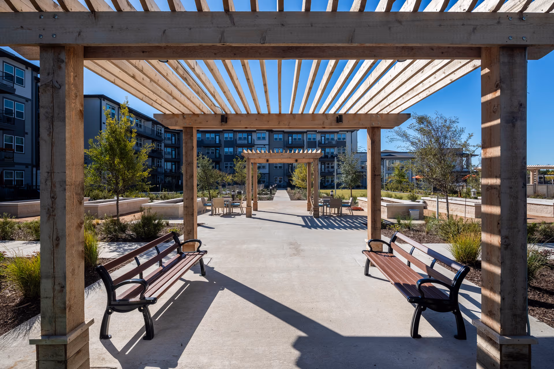 Outdoor seating area at Amberlin at The Station featuring wooden pergolas casting shadows on a concrete pathway with benches on either side. There are trees and landscaped garden beds surrounding the area, with multi-story residential buildings in the background under a clear blue sky.