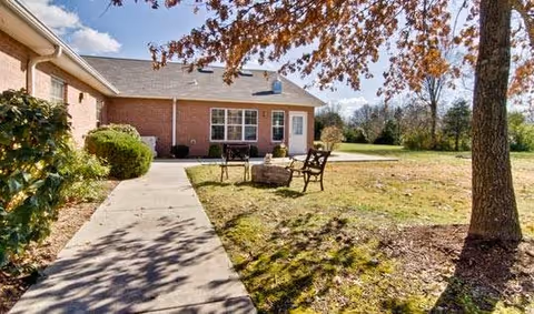 Outdoor view of a brick building with a concrete walkway leading to a door. There are two benches and a fire pit on a grassy area next to the walkway. Trees and bushes surround the area under a partly cloudy sky.