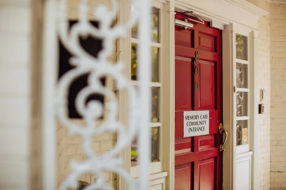 A red door with the number 800 and a sign that reads 'Memory Care Community Entrance' on it, surrounded by white brick walls and windows with white frames.