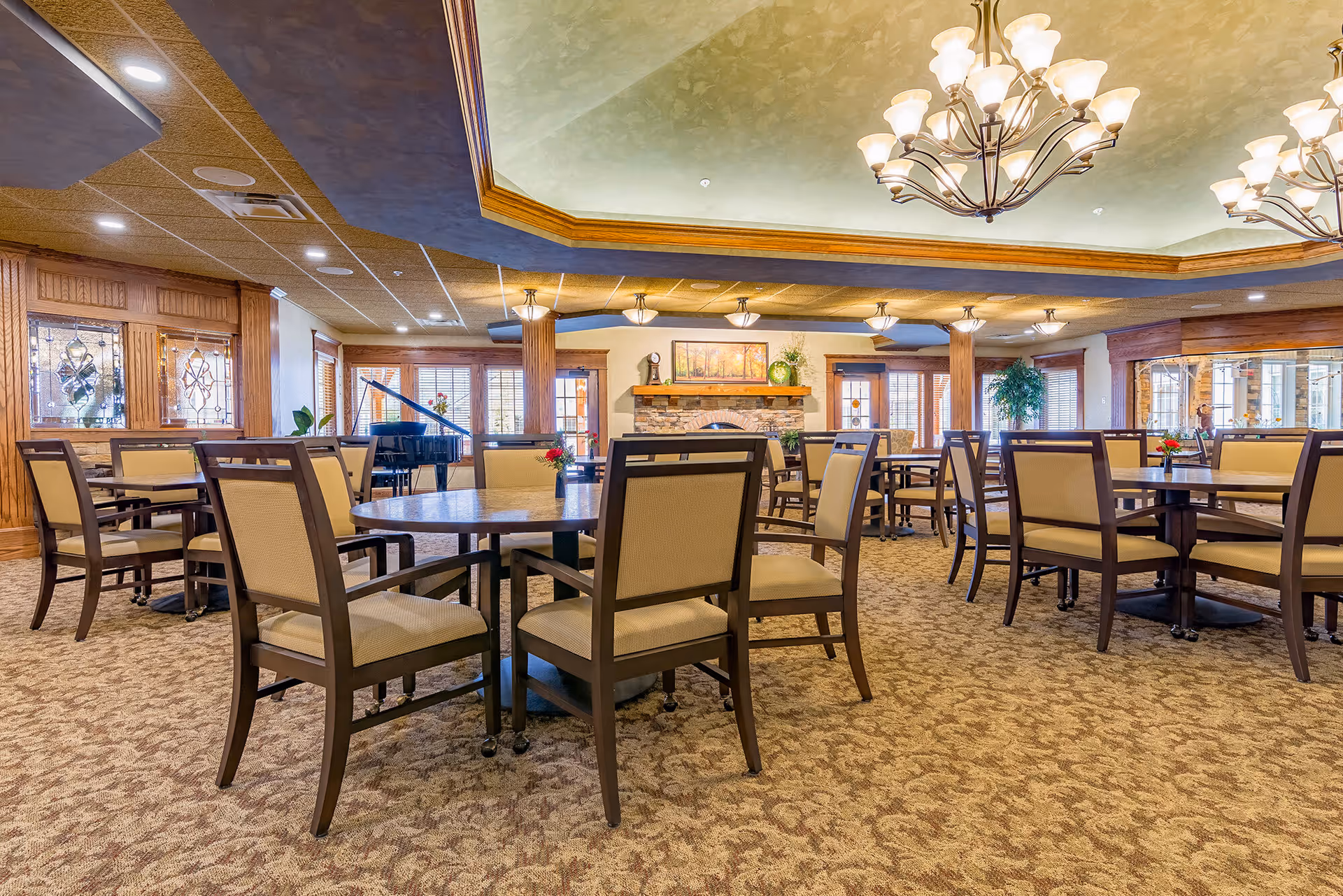 Spacious dining room with multiple round tables and cushioned chairs arranged neatly on a patterned carpet. The room features warm lighting from chandeliers and ceiling lights, a stone fireplace with a painting above it, large windows with wooden blinds, and a black grand piano in the corner.